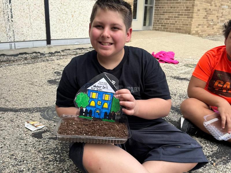A boy proudly holds a decorated house model made from paper in a planting container.