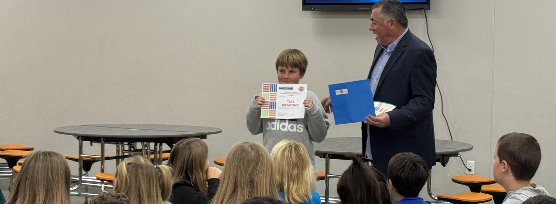 A boy holds a certificate while a man stands beside him in front of an audience.