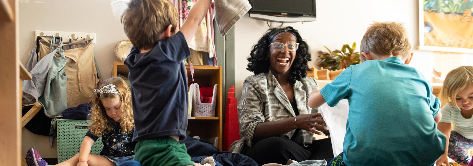 Children playing with fabric and a smiling teacher in a classroom.