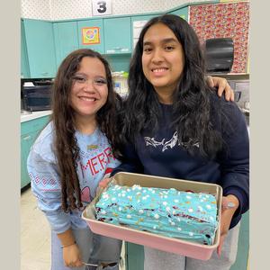 Two students proudly hold a blue-frosted cake in the kitchen.