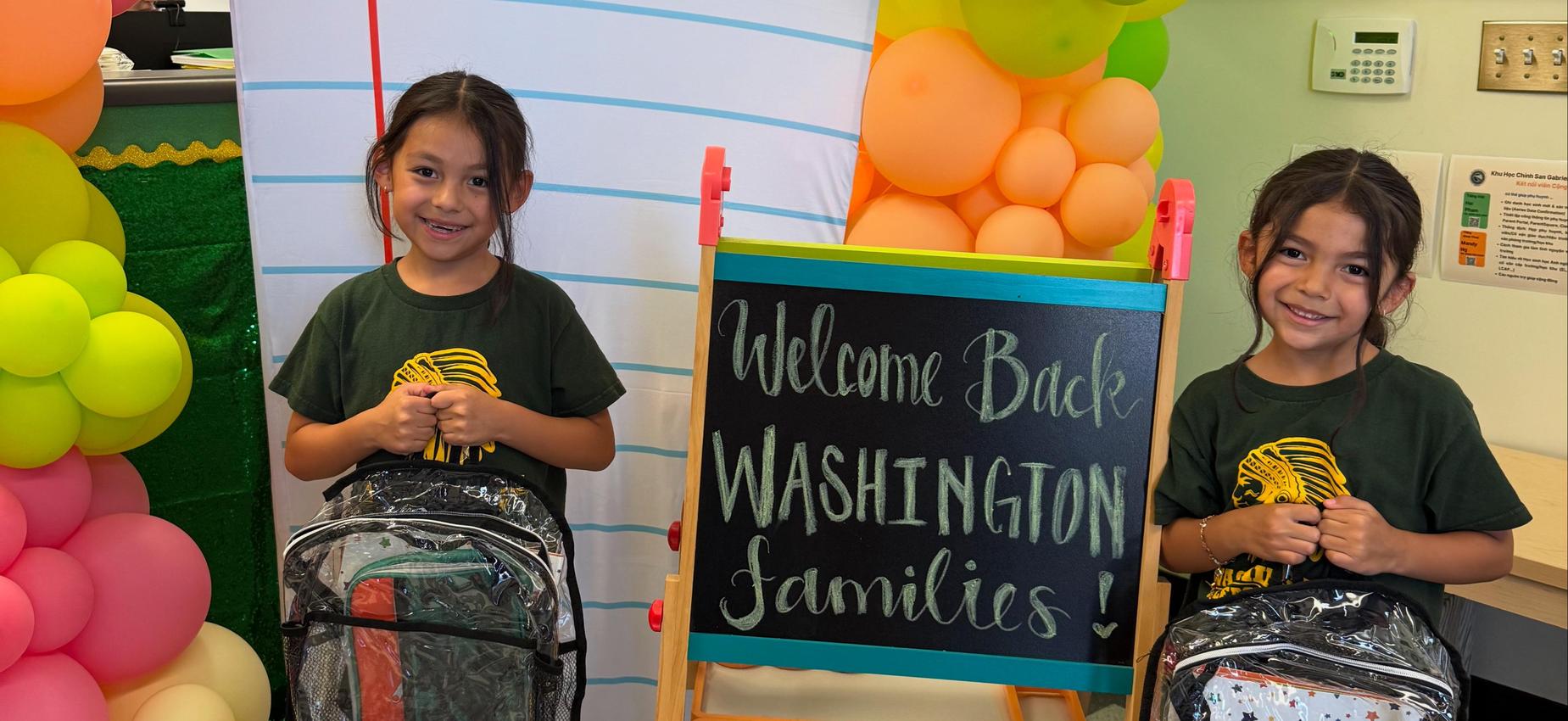 Two girls in green shirts with a welcome sign and colorful balloons.