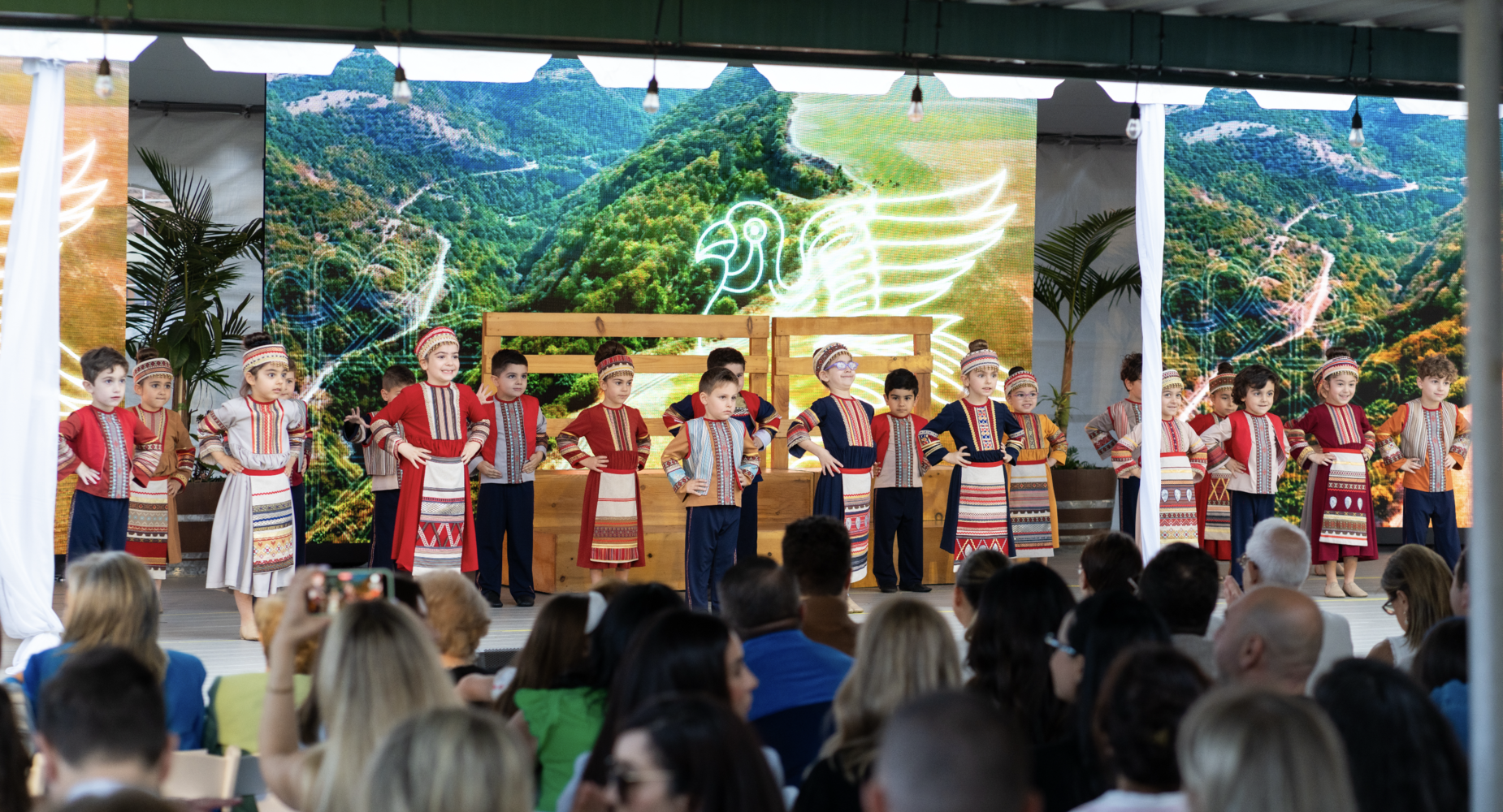 Children performing on stage with traditional costumes in front of a scenic backdrop.