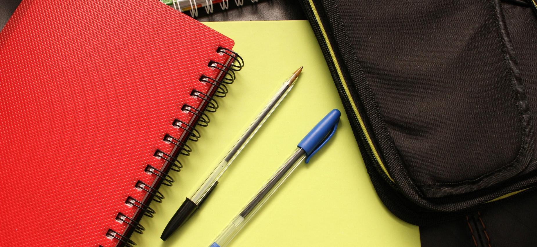 Red notebook, pens, and a pencil case on a desk.
