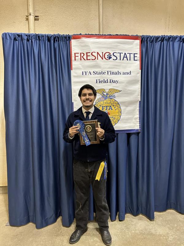 A male FFA student stands alone in front of the “Fresno State FFA State Finals and Field Day” banner, smiling and holding a first-place blue ribbon and a plaque recognizing his achievement.