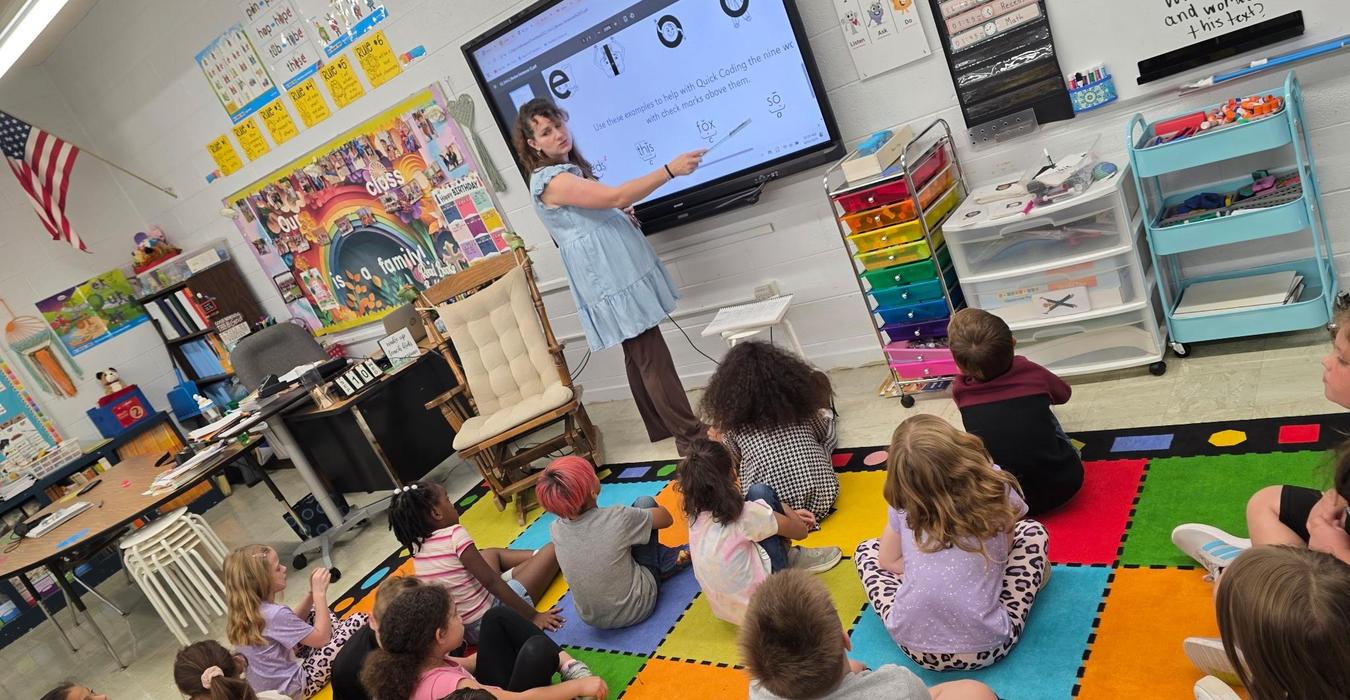 A teacher instructs a group of young students sitting on a colorful carpet in a classroom.