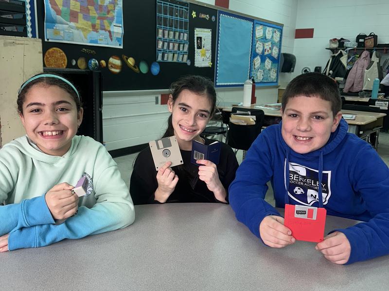 Three smiling children holding up floppy disks in a classroom setting.