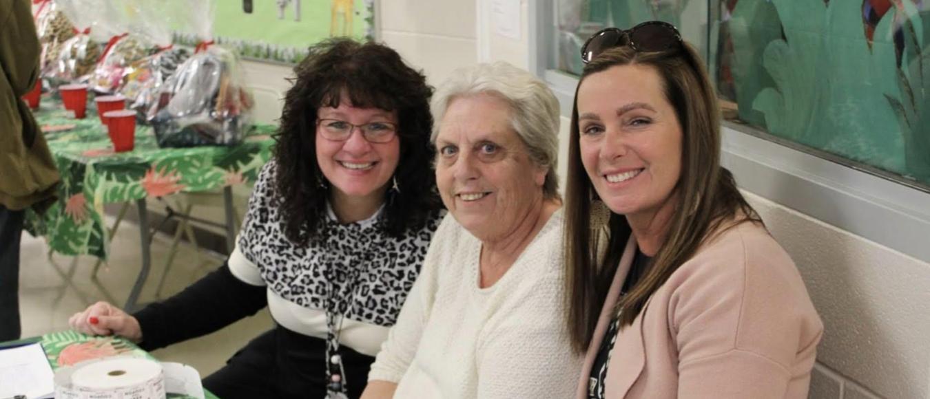 Three smiling women sit together behind a green patterned table with a roll of raffle tickets.