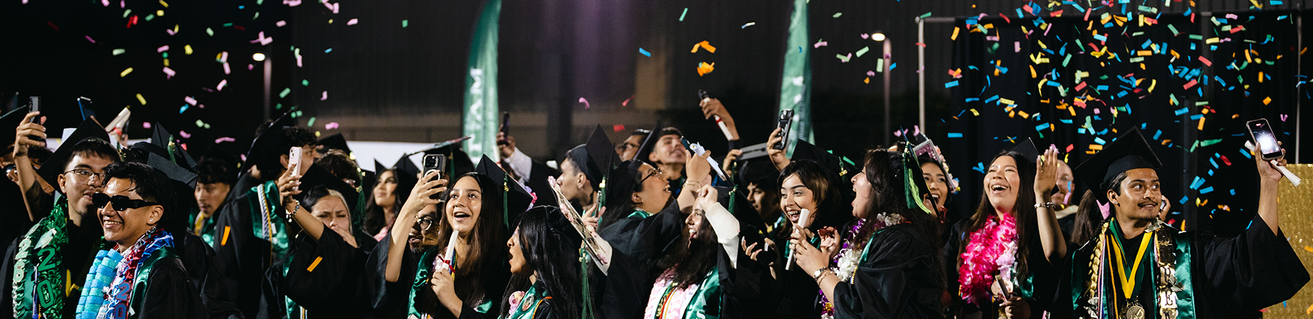 A large group of high school graduates wearing caps and gowns celebrate with confetti overhead