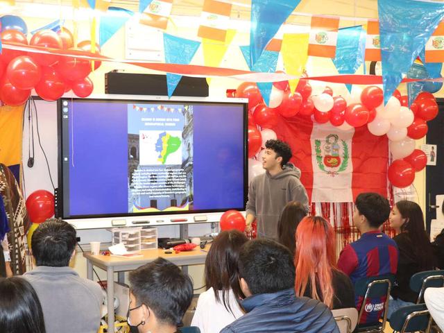A presenter stands before a screen displaying a map, surrounded by balloons and flags.