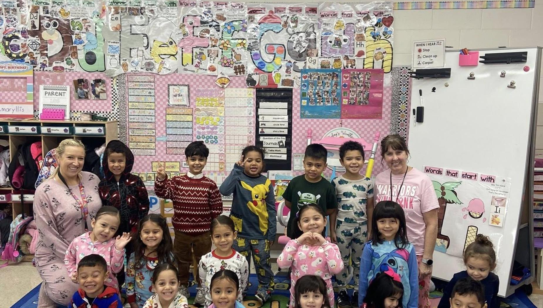 Large group of children and adults, some in festive pajamas, in a decorated classroom.