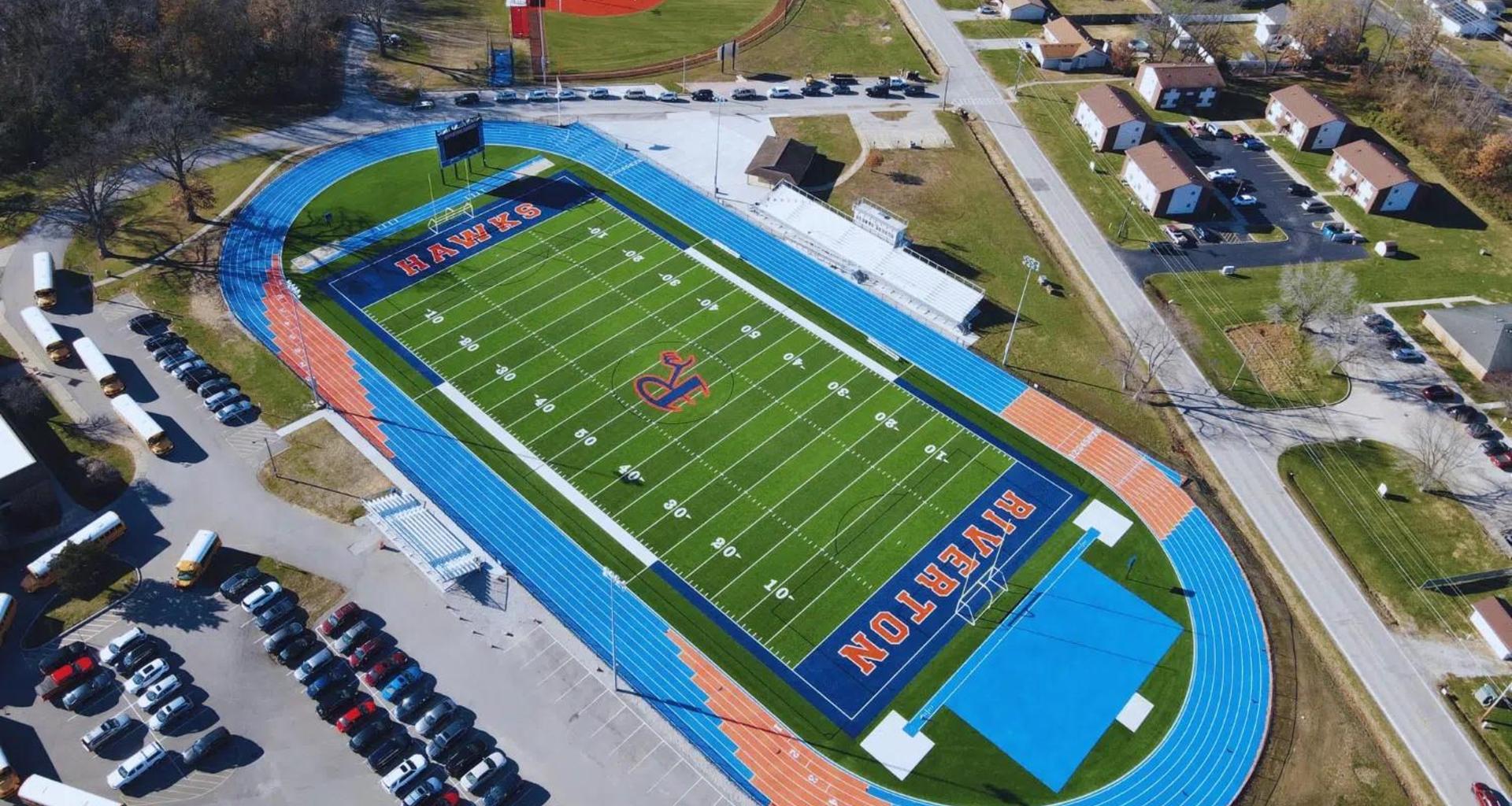 Aerial view of a sports field with blue track and surrounding buildings.