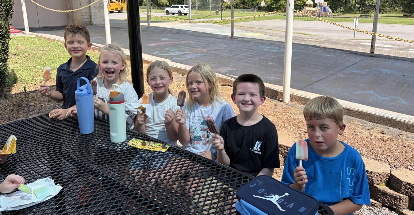 Group of children sitting at a table, holding ice pops and smiling.
