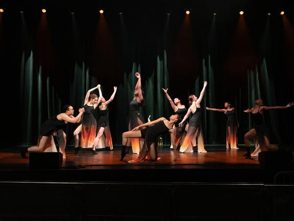 A group of dancers in black costumes performing on stage with dramatic lighting.