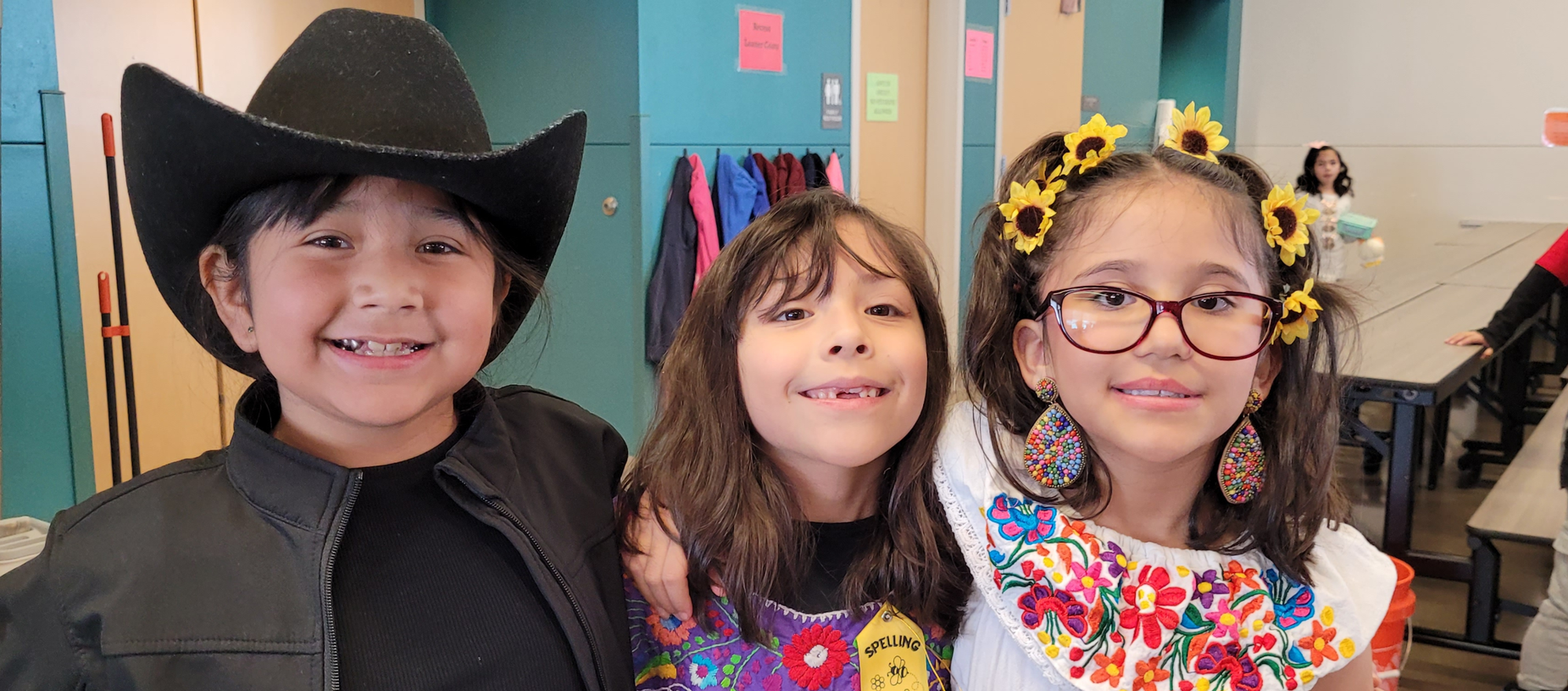 Three children smiling, dressed in colorful attire with accessories, posing together.
