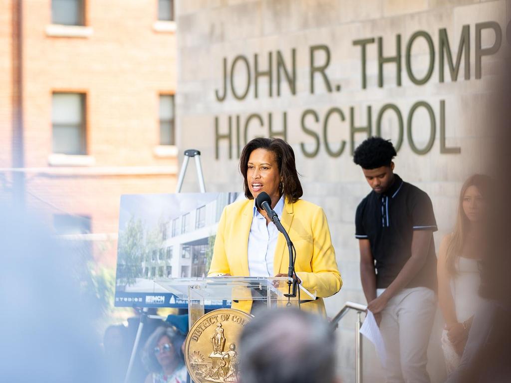 A woman in a yellow jacket speaking at a podium with students and a school backdrop.