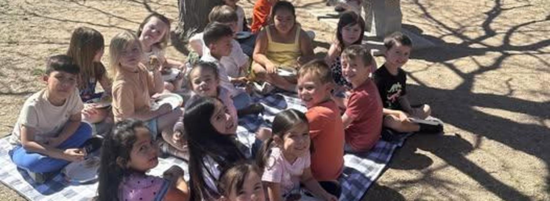 A group of children sitting on a picnic blanket under a tree, enjoying a meal together.