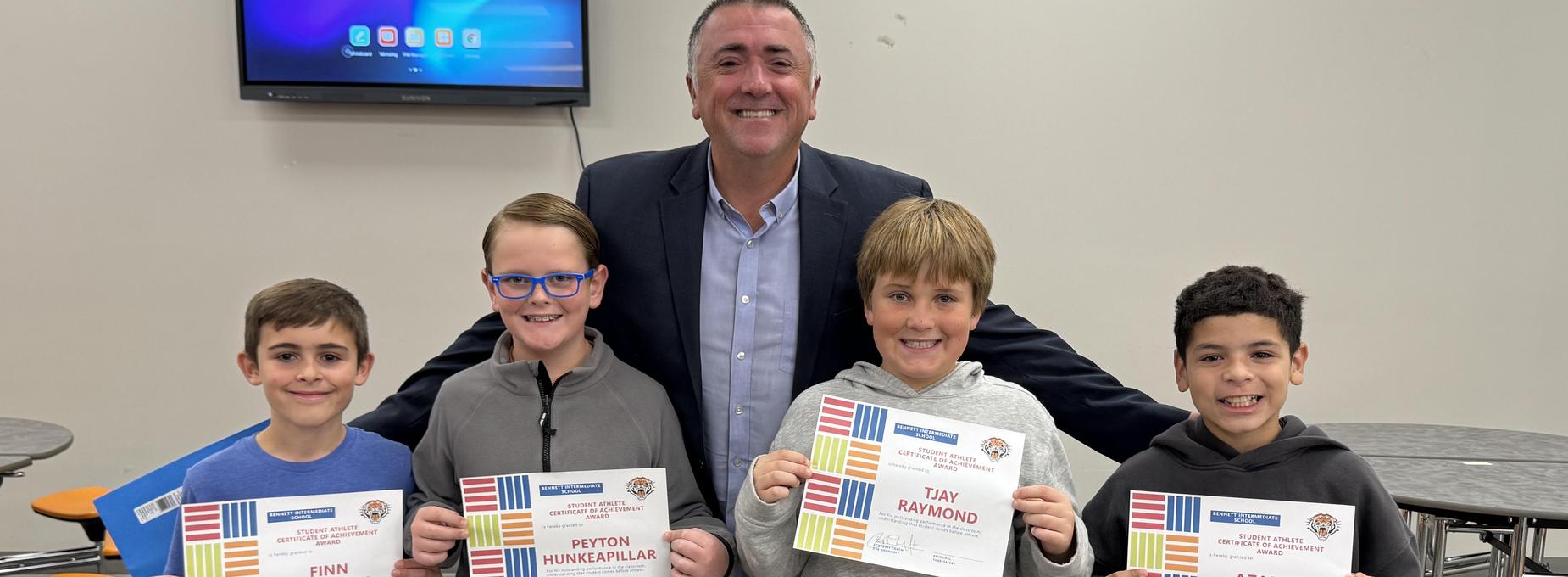 Four boys hold certificates, smiling with a man in a blazer behind them.