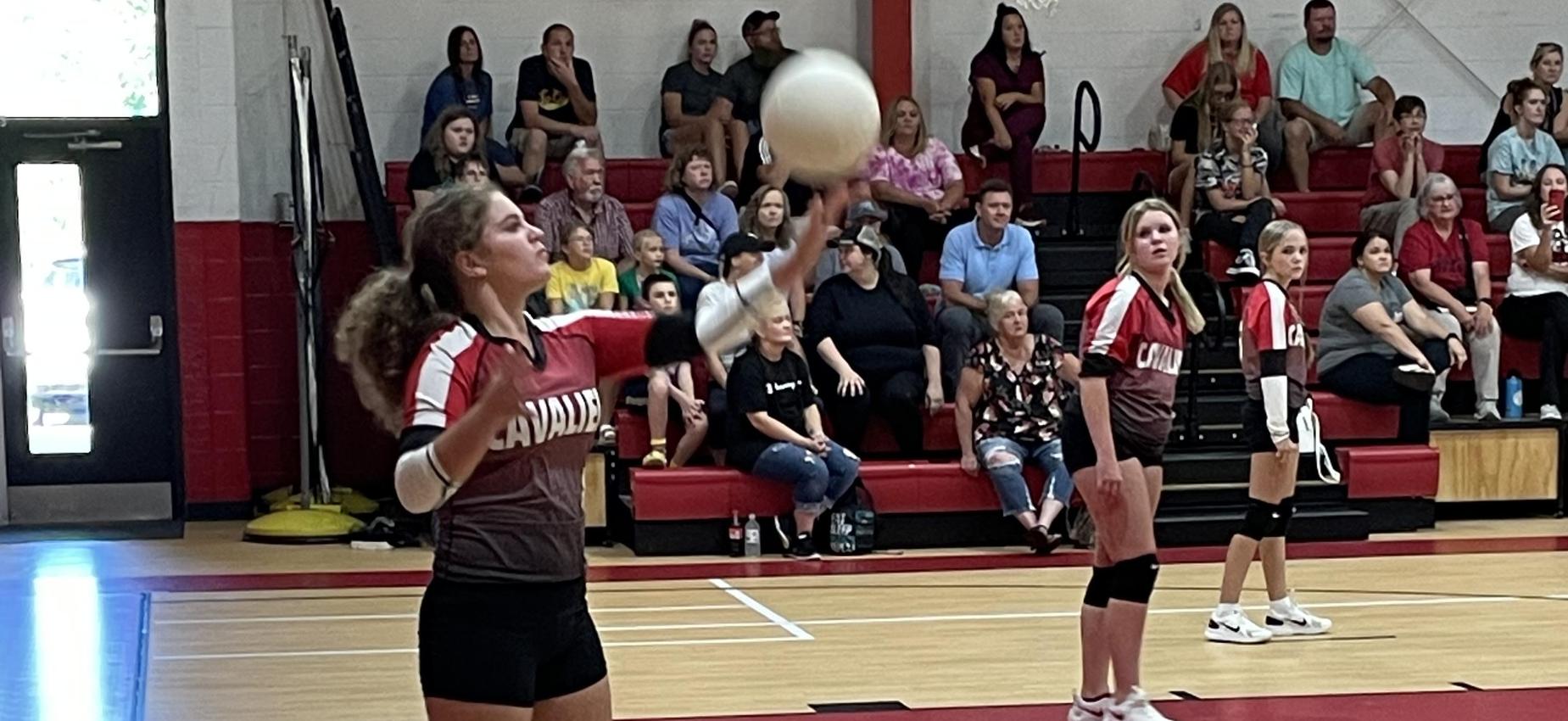 A volleyball player serves the ball while two players look on.