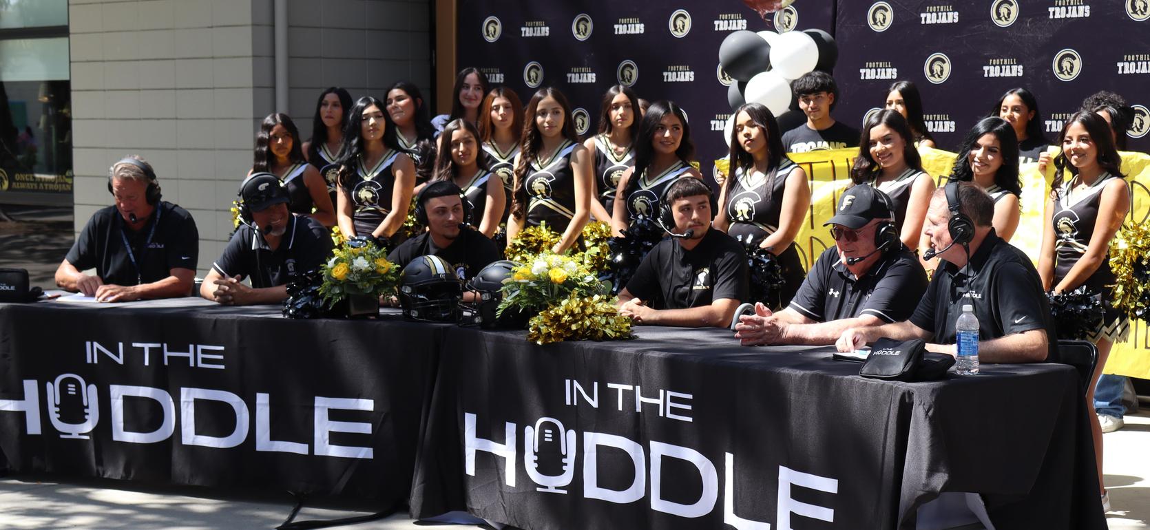 Five commentators at a table with a cheerleading team in the background.