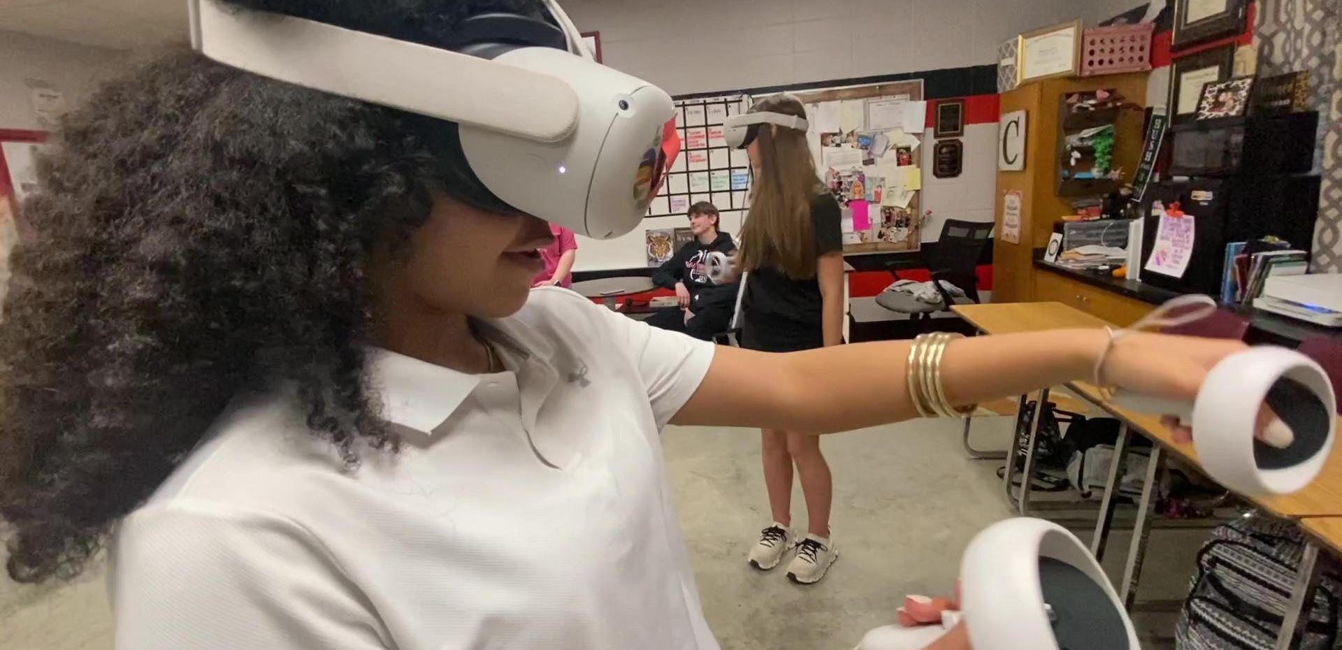 A girl wearing a VR headset interacts with controllers in a classroom setting.