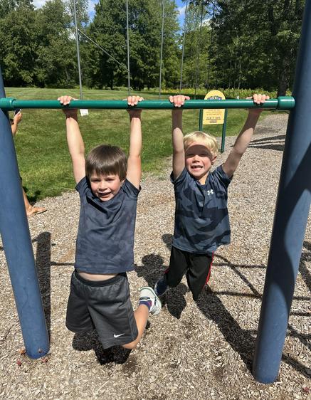 Two boys hang from a playground bar, showing excitement in a green park.