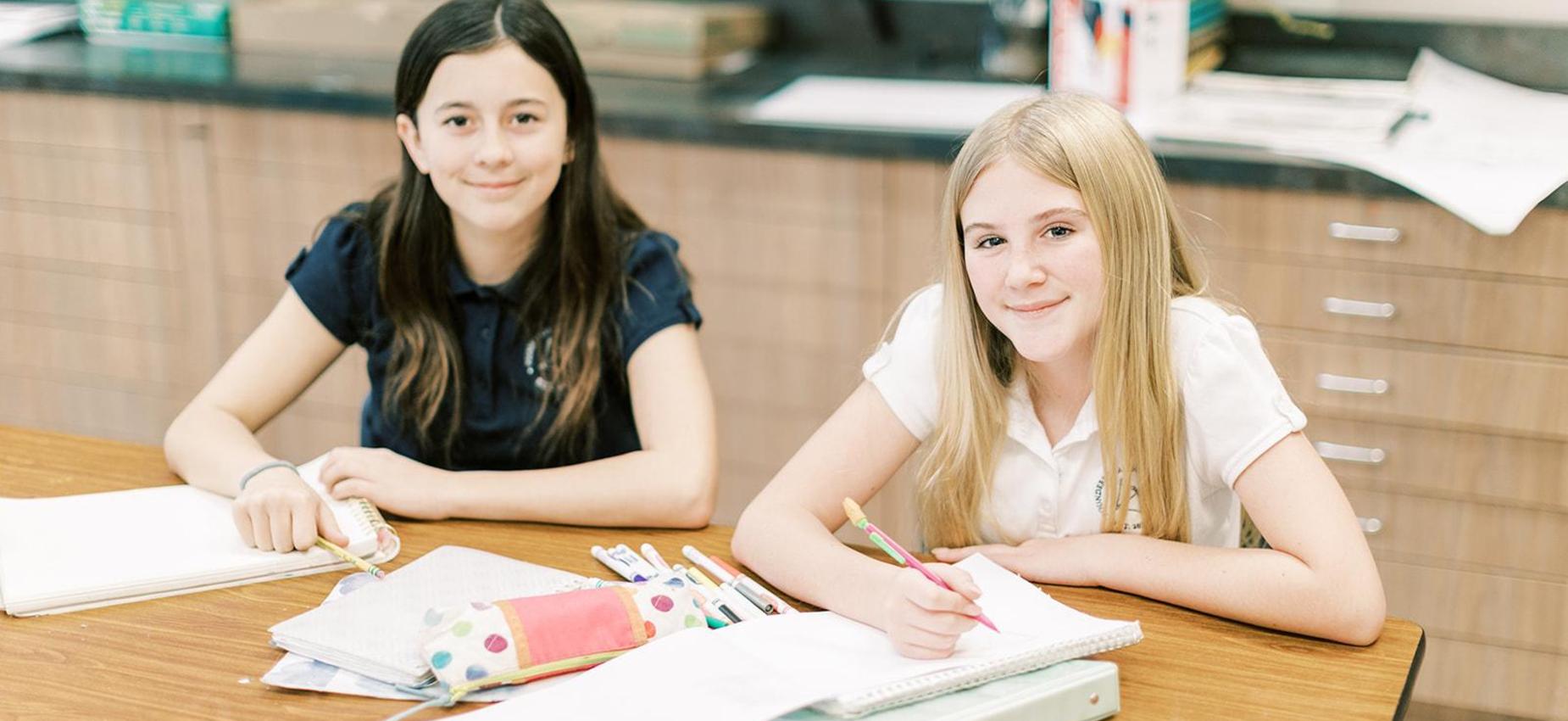 Two smiling middle school girls draw at a classroom table
