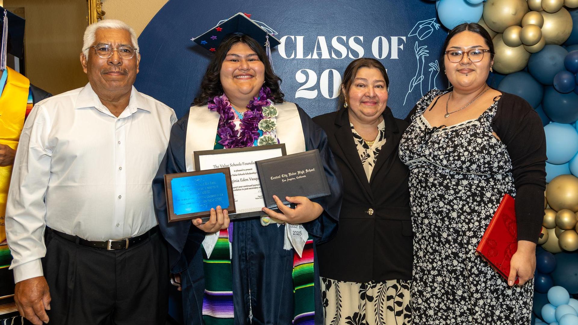 A graduate poses with family holding diplomas and awards at a graduation celebration.