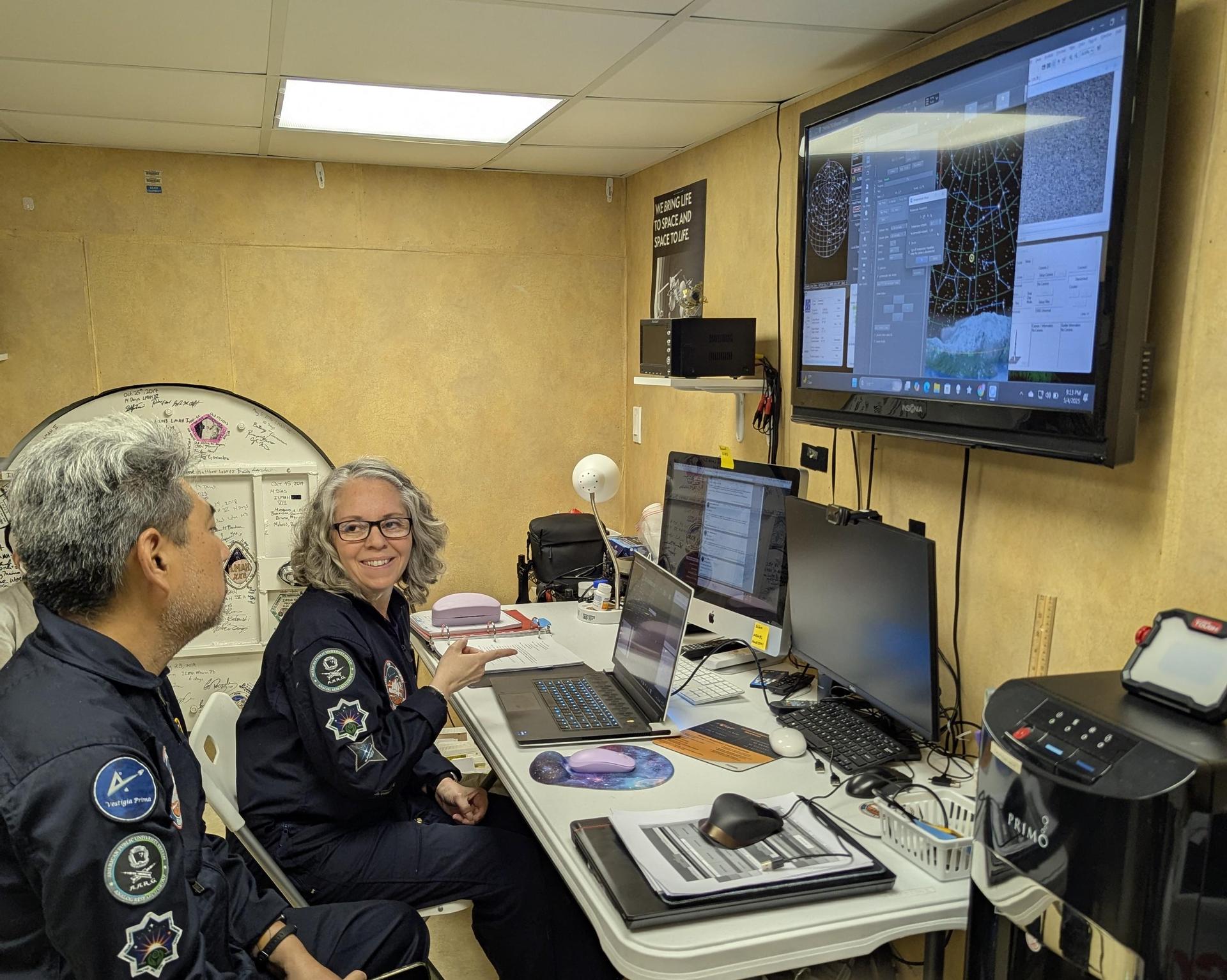 Two people in flight suits working at computers in a control room.