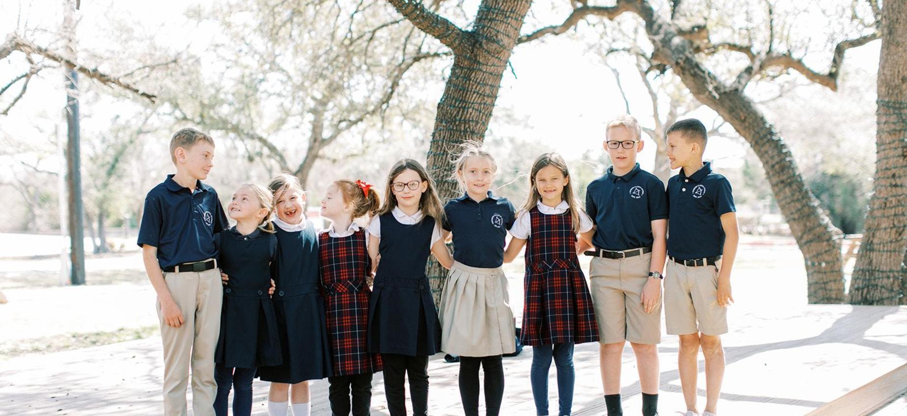 Elementary and middle school students in uniform stand in a row under oak trees