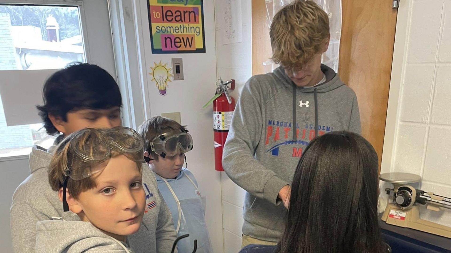 Boys in gray sweatshirts observe a teacher during a hands-on science activity.