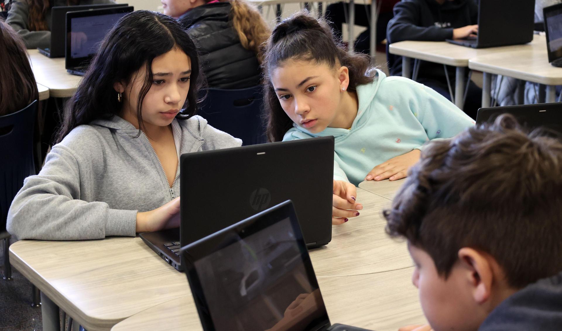 Girl leans over to another girl to help with her work