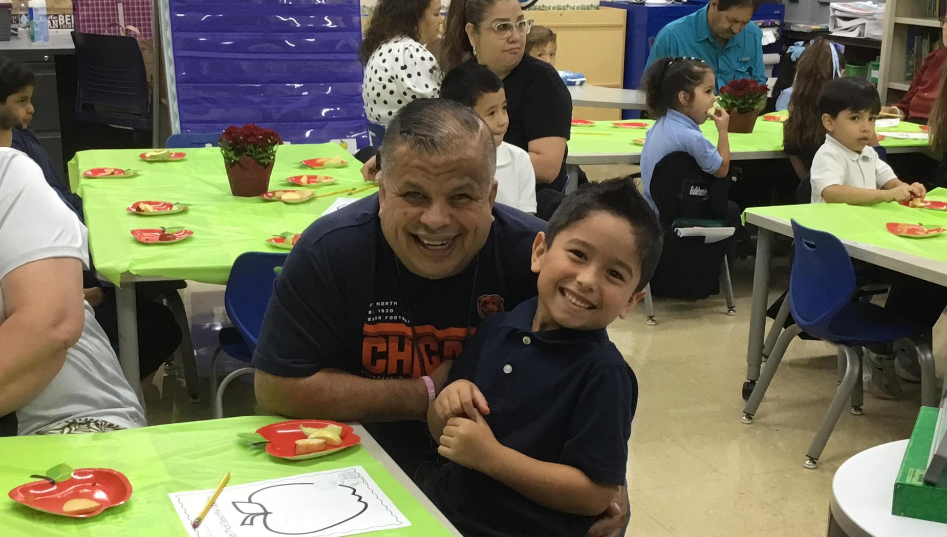 A smiling boy sits with an older man in a classroom filled with children and snacks.