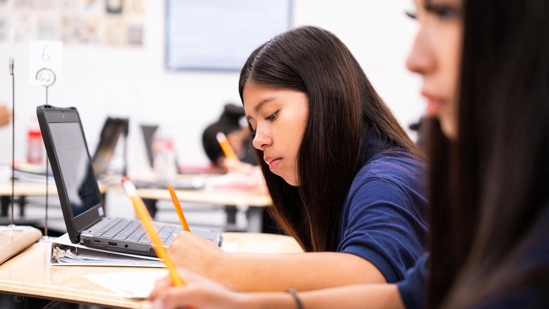 Student focused on writing notes beside a laptop in a classroom setting.