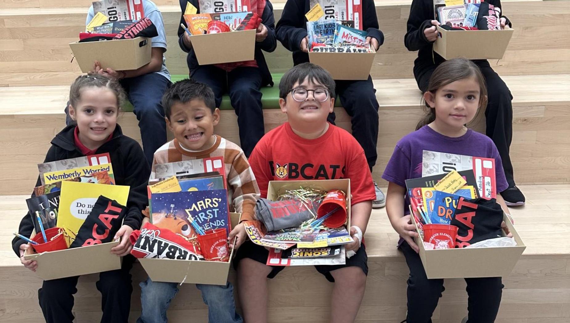 Children sitting on steps, each holding a box of books and school supplies.