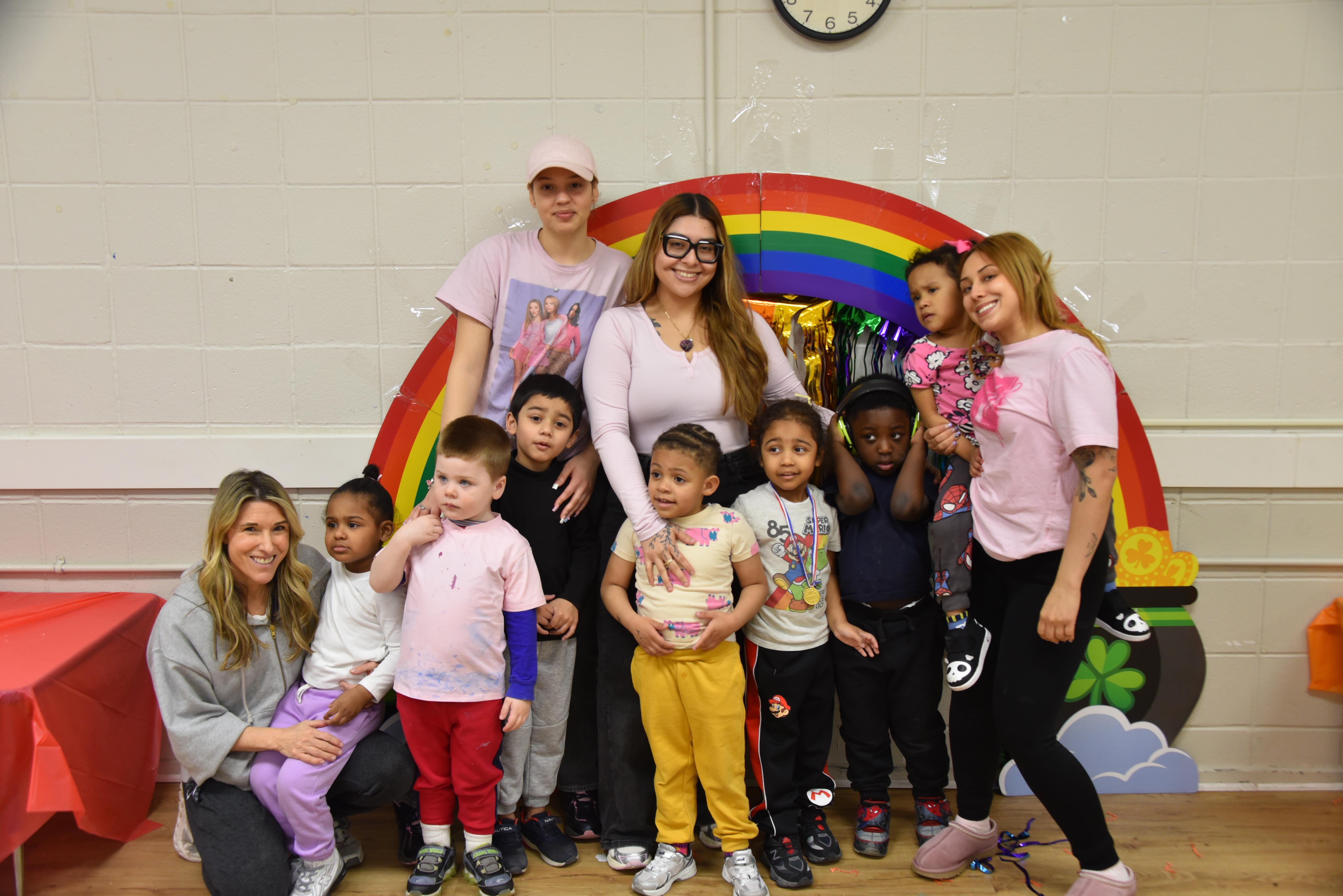 3 teachers and a class of preschoolers are standing in front of a rainbow