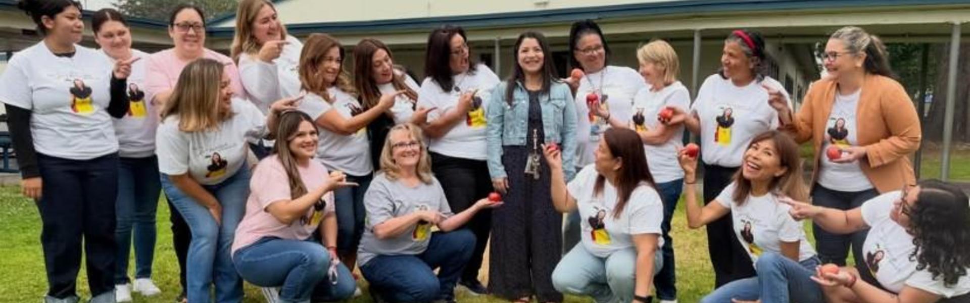 Group of smiling women in matching t-shirts holding apples outdoors.