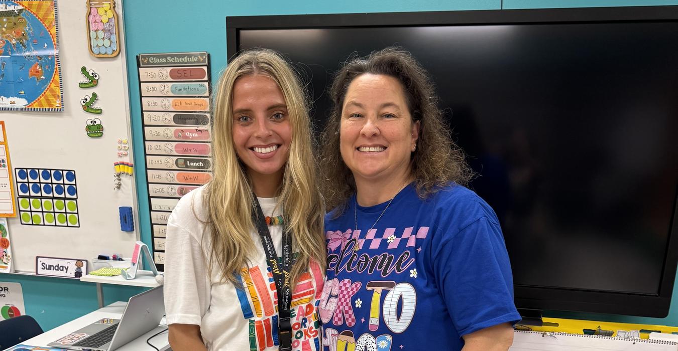 Two women smiling together in a classroom setting with colorful decor.