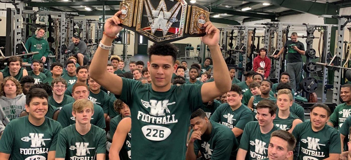 large group of football players kneeling together in the weightroom. one player is standing with a weight belt held above his head.