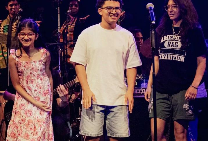 Three young people stand on stage smiling, with a band in the background.