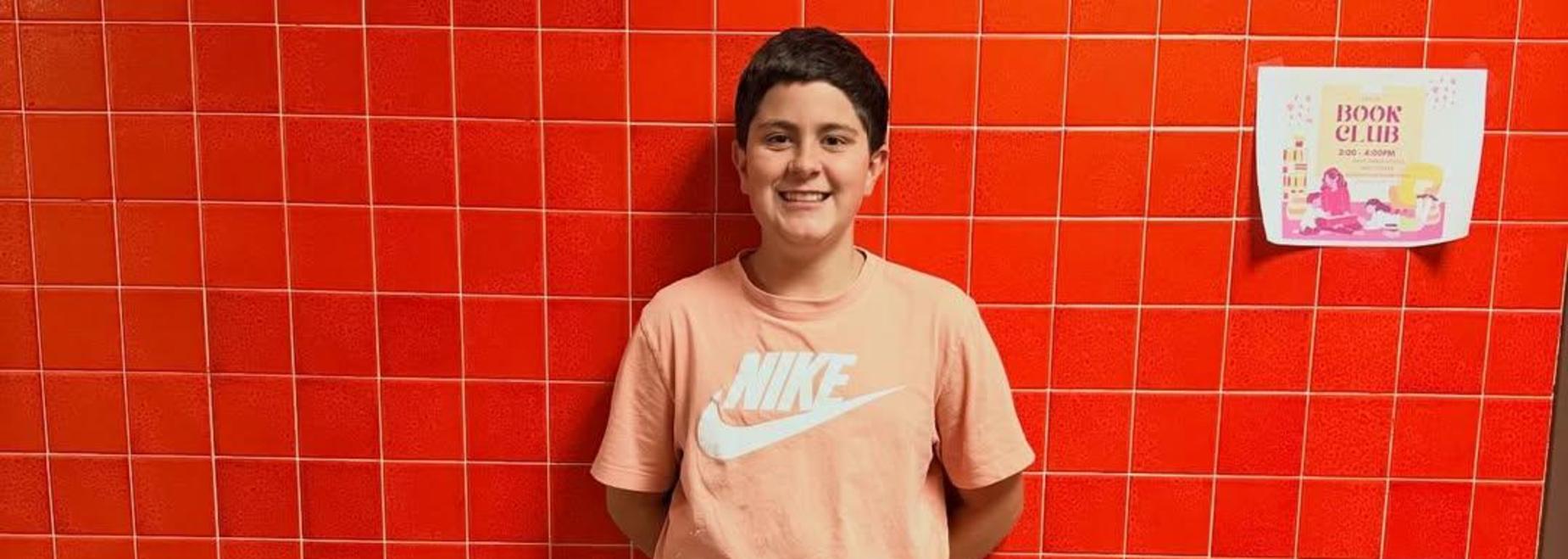 Child standing against a bright red tiled wall, wearing a pink Nike shirt and black pants.