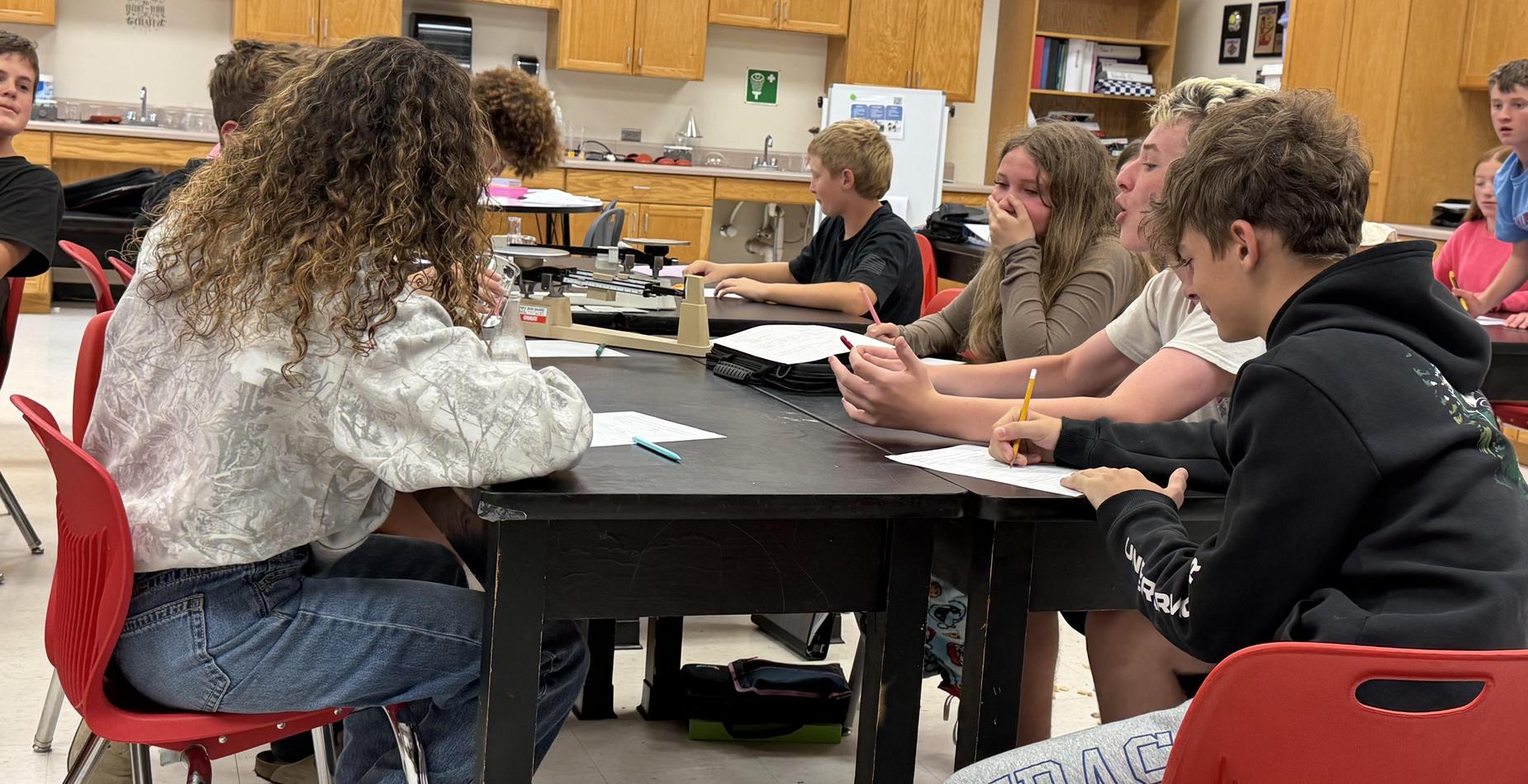 Group of students engaged in discussion and note-taking around a classroom table.