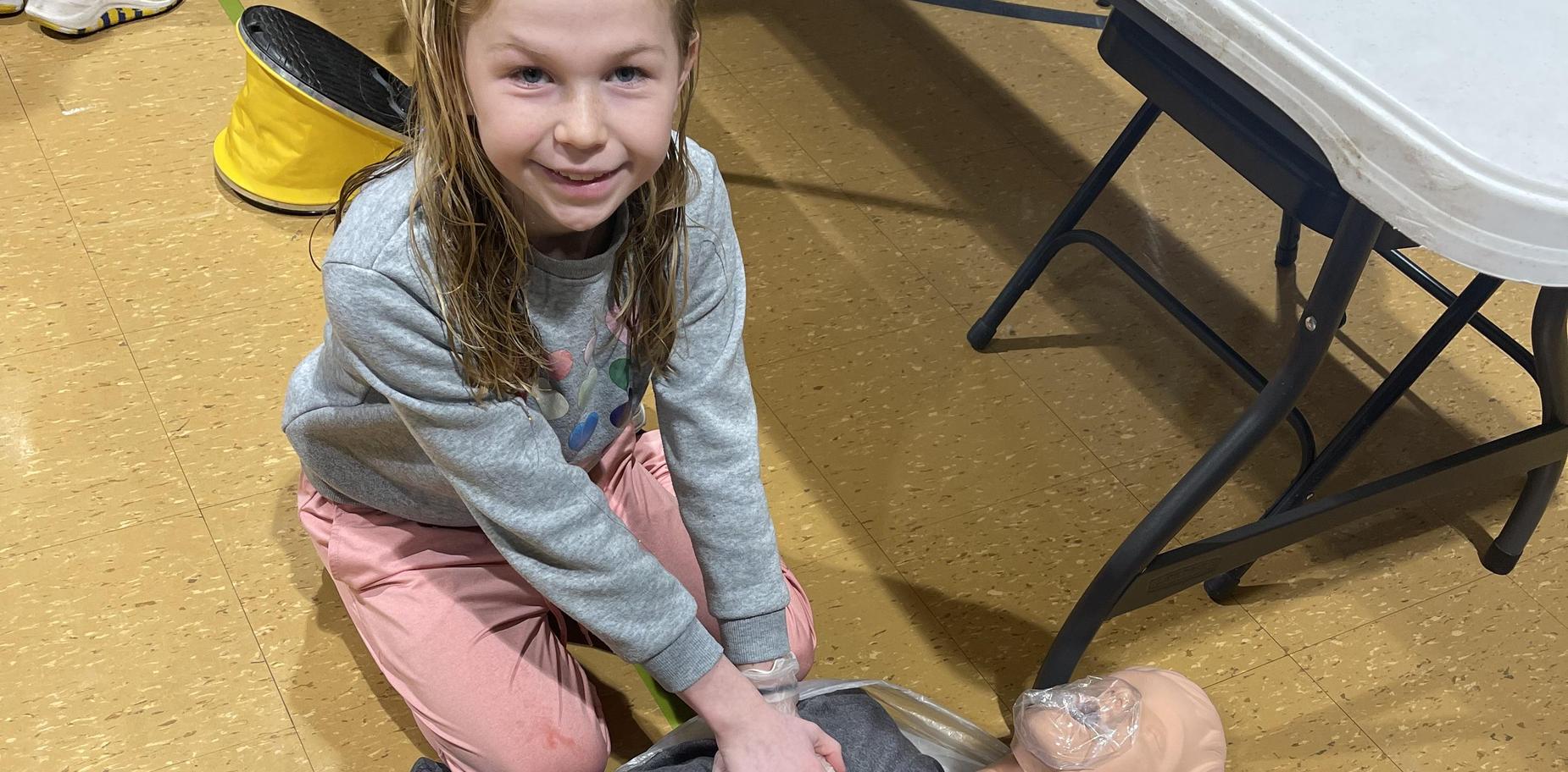Girl practicing CPR on a training dummy while sitting on a floor covered with a plastic sheet.