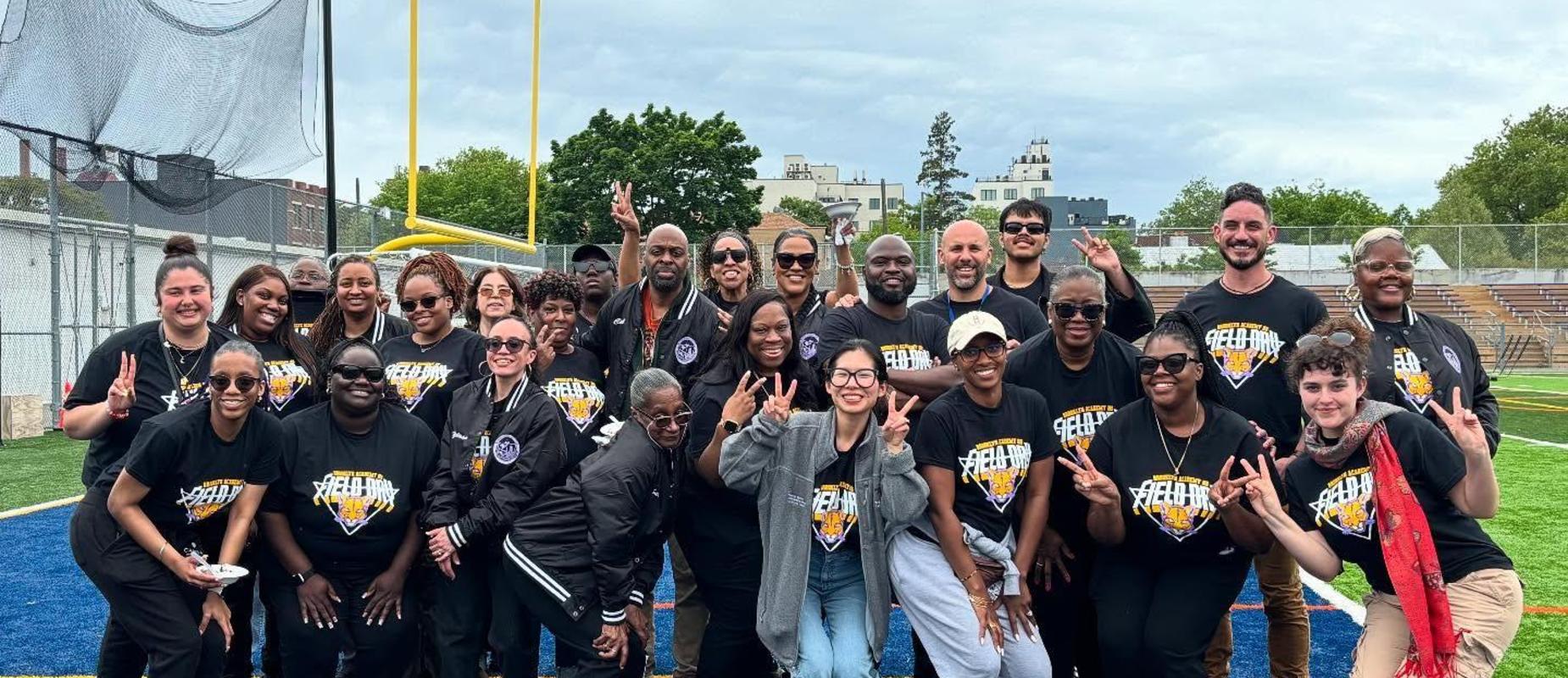 Large group of people posing together on a sports field, smiling and making hand signs.
