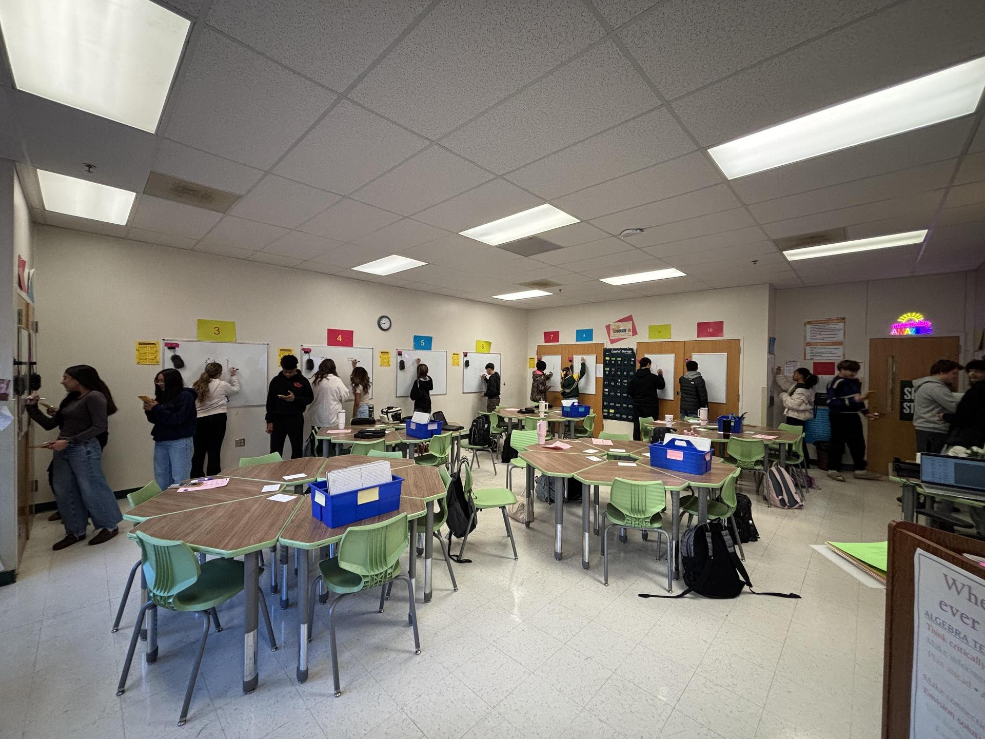 A classroom filled with students standing and working on tasks at various tables.
