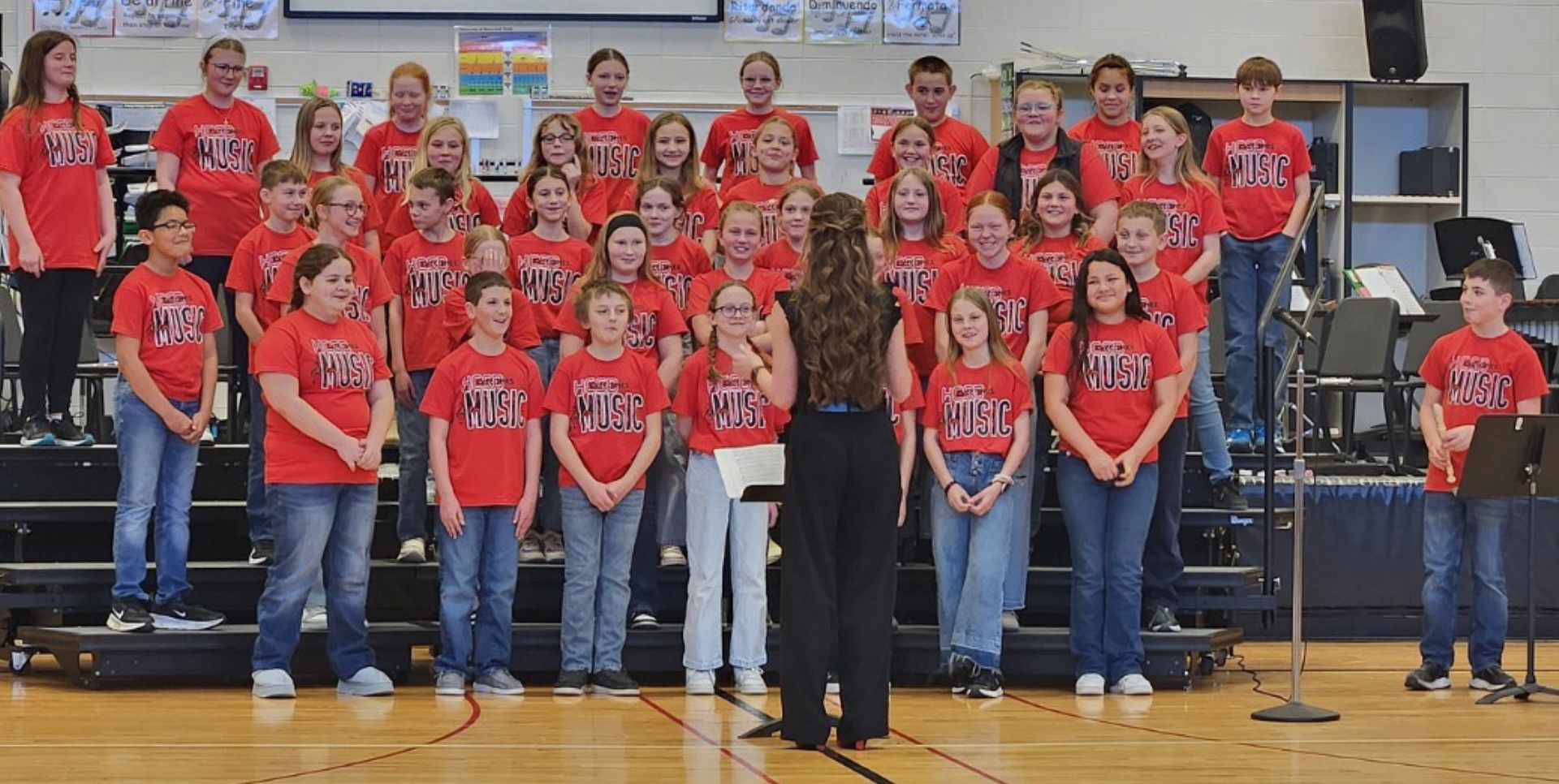 Chorus group of students in red shirts performing on a stage with a conductor.