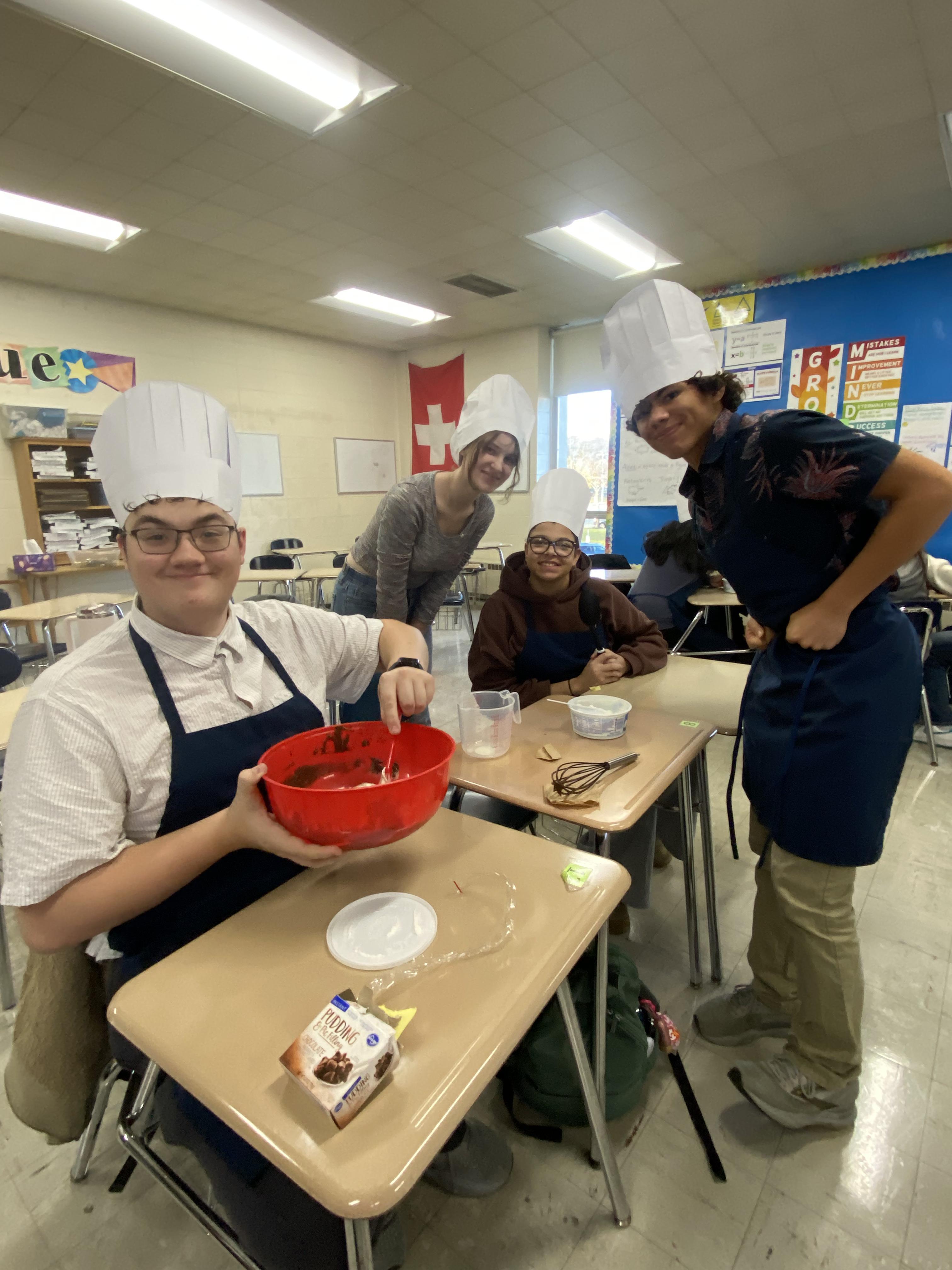 Students in chef hats preparing food at their desks with smiles.