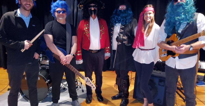 A group of five musicians in costumes with colorful wigs and instruments on stage.