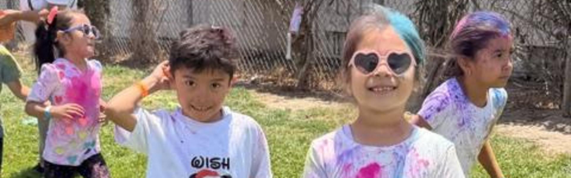 Two children stand on grass, wearing colorful shirts, with others playing in the background.