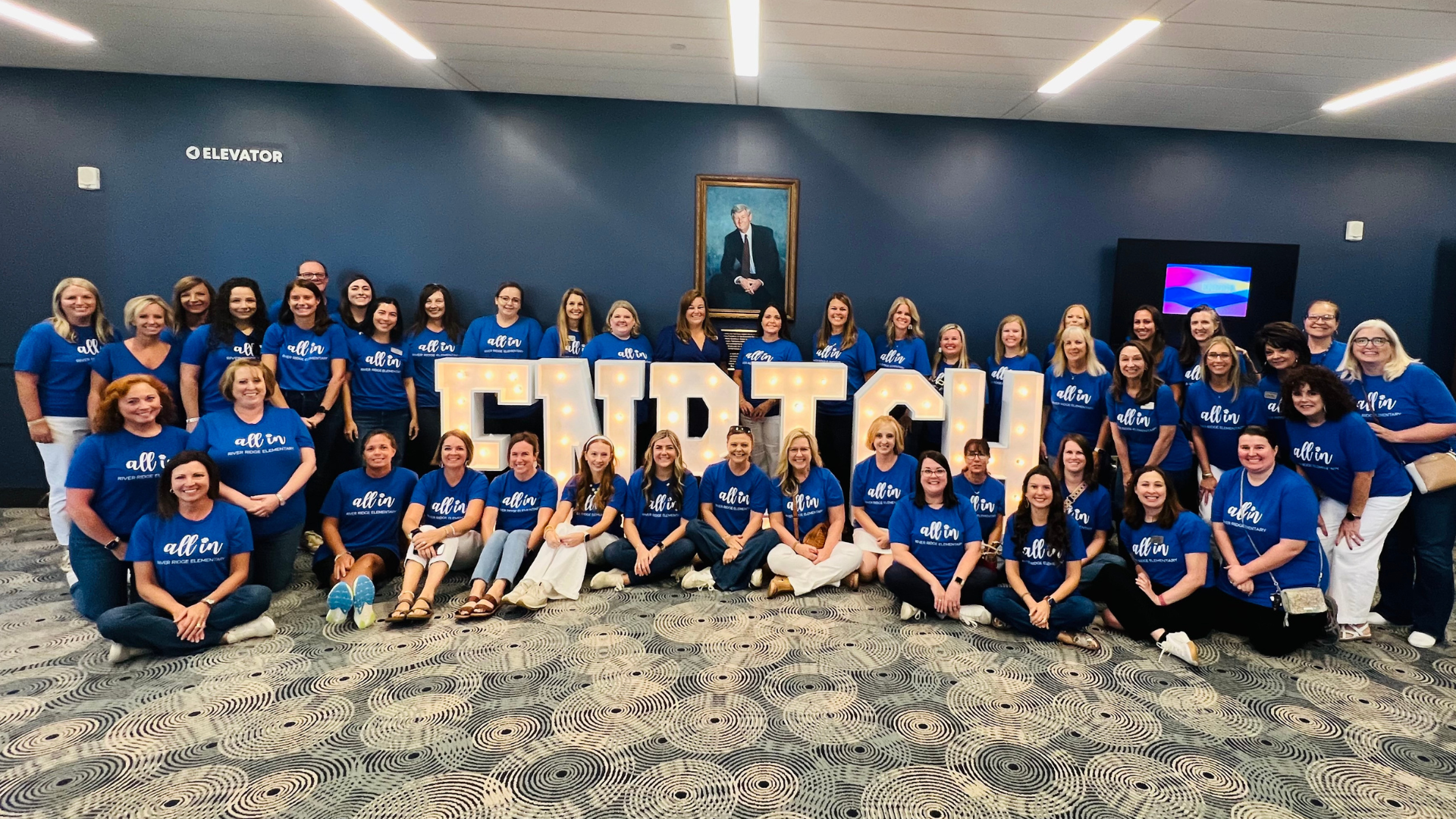 Group of women in blue shirts posing together in front of a large illuminated sign.