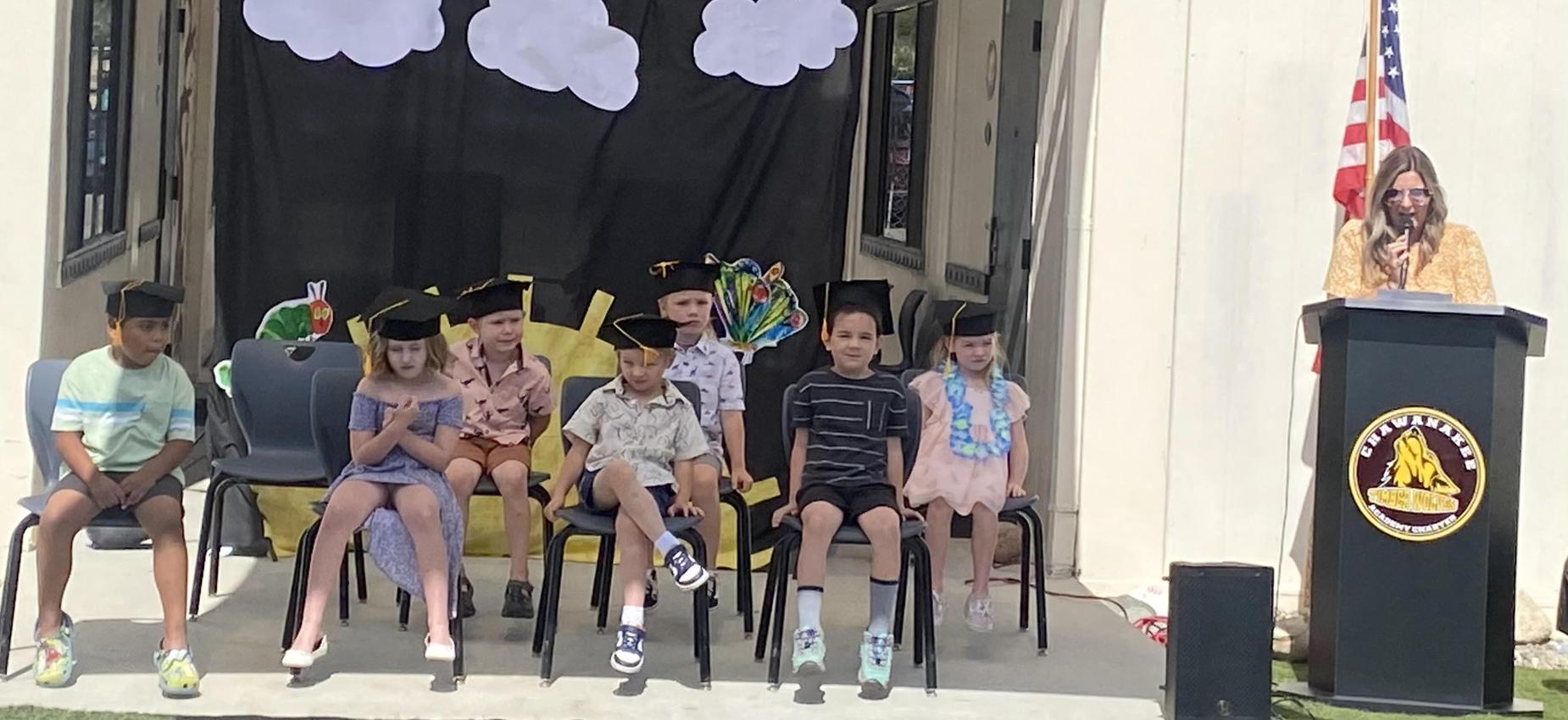 Graduating children in caps sitting on stage with a speaker and decorations.